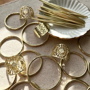 May include: A collection of gold-toned metal rings, bangles, and decorative pieces. The items are arranged on a beige fabric surface. Some of the pieces have a textured finish, while others have a delicate, openwork design.