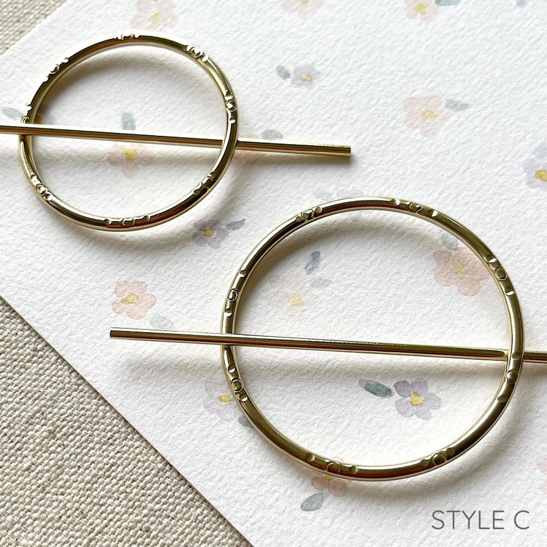 May include: Two gold metal hair clips with a circular design and a straight bar across the centre. The clips are on a white background with a floral pattern.