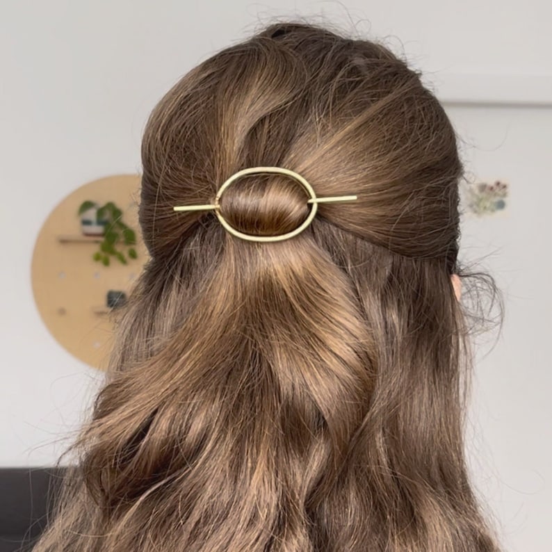 May include: A gold oval-shaped hair clip with a straight bar across the center. The clip is in a woman's brown hair.