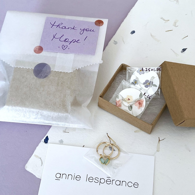 May include: A small package with a handwritten thank you note that says "Thank you Hope!" and a heart. The package is sitting on a table with a white background. There are two small boxes with jewelry inside. One box has a butterfly charm necklace and the other has a delicate chain necklace with a small charm. The jewelry is packaged in clear plastic bags. The boxes are made of brown cardboard. The table is covered in white paper with small, colorful confetti scattered on it. The image also includes a card with the text "annie lesp&egrave;rance" written on it.