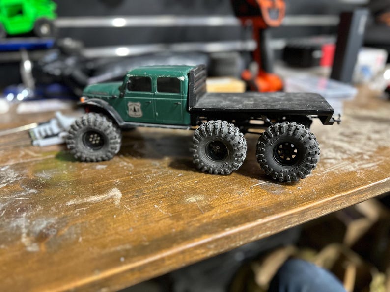 Peut inclure: Un camion jouet vert avec une benne et quatre grandes roues noires. Le camion a une cabine noire avec des fen&ecirc;tres et un pare-chocs noir. Le camion est pos&eacute; sur une surface en bois.