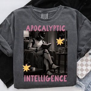 Op de afbeelding: Een antracietgrijs t-shirt met een grafische print van een witte robot die in een fauteuil zit en een boek leest. De tekst "APOCALYPTIC" staat in het roze boven de afbeelding en "INTELLIGENCE" in het roze eronder, met gele sterpictogrammen.