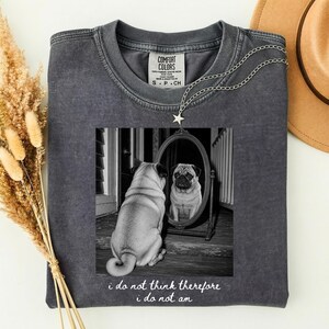 May include: A charcoal gray crewneck t-shirt featuring a black and white graphic of a pug sitting in front of an oval mirror, with the text "I do not think therefore I do not am" printed below the image. The shirt has a vintage washed appearance.