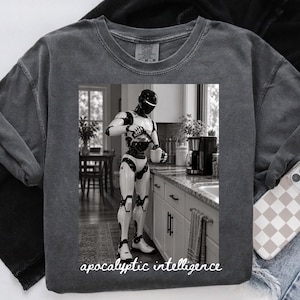 Op de afbeelding: Een antracietgrijs t-shirt met een zwart-witte afbeelding van een slanke, humanoïde robot die koffie inschenkt in een keuken. De robot heeft een gladde, glanzende afwerking en staat bij een aanrecht. De tekst "apocalyptic intelligence" staat eronder gedrukt.
