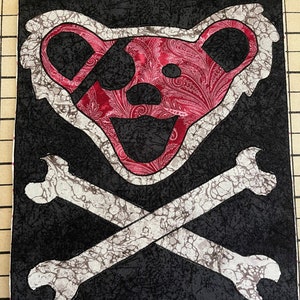 May include: A black and white fabric patch with a red and white paisley bear head with a black eye patch and crossed bones. The bear head and bones are outlined in white.