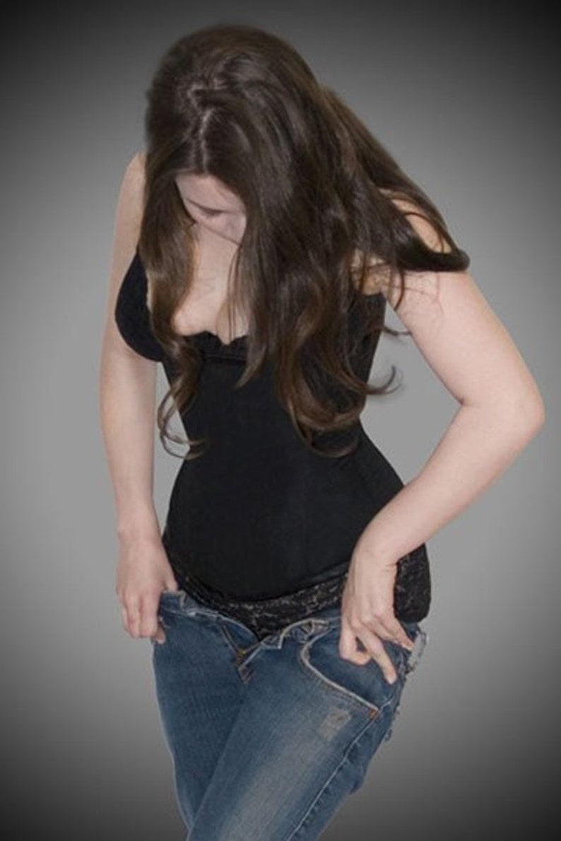 Pode incluir: Um top preto estilo espartilho com acabamento em renda &eacute; combinado com jeans azuis. A pessoa est&aacute; ajustando os jeans, com as m&atilde;os nos bolsos. O fundo &eacute; um gradiente de cinza.