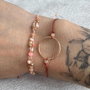 Peut inclure: Deux bracelets sur un poignet. Un bracelet est fait de ficelle rouge avec un charme rond en or. L'autre bracelet est fait de perles roses et dorées.