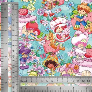 May include: A patterned fabric featuring various cartoon characters, strawberries, and flowers on a light blue background. A metal ruler is visible along the left side and bottom edge, showing measurements in both inches and centimeters.