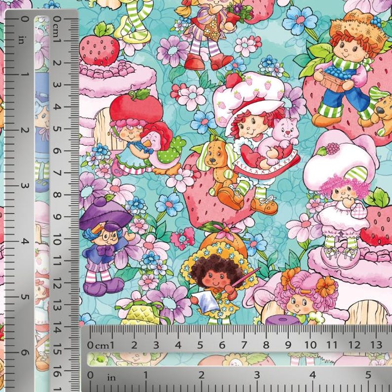 May include: A patterned fabric featuring various cartoon characters, strawberries, and flowers on a light blue background. A metal ruler is visible along the left side and bottom edge, showing measurements in both inches and centimeters.