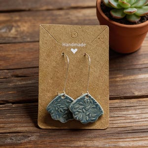 May include: A pair of handmade blue ceramic earrings with a leaf pattern, displayed on a brown card with the word "Handmade" and a heart. The earrings hang from silver hooks. A small potted succulent is in the background.