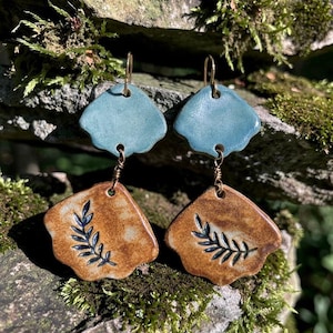 May include: Handmade ceramic earrings. Each earring features a light blue, scalloped top piece and a brown, rectangular bottom piece with a dark blue leaf design. The earrings are connected with gold-colored wire and hang from gold-colored ear hooks.