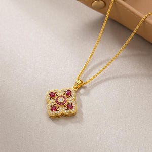 May include: A gold chain necklace featuring a square-shaped pendant with rounded corners. The pendant is decorated with four pear-shaped red stones, a central round white stone, and small white accent stones set in a gold filigree frame.