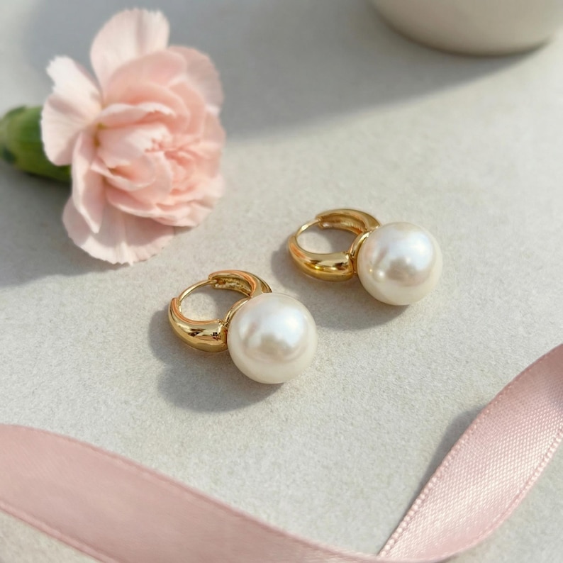 Puede incluir: Un par de pendientes de aro dorados con grandes perlas blancas redondas unidas a la parte inferior de cada aro. Los pendientes se muestran junto a un clavel rosa claro y una cinta de color rosa claro a juego.