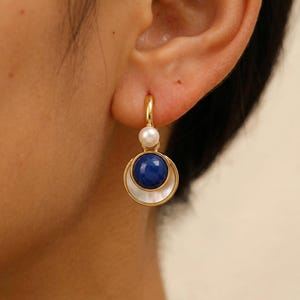 Può includere: Orecchini a cerchio dorati con una perla e una pietra blu. Gli orecchini presentano una piccola perla bianca in alto, una pietra blu scuro rotonda e un accento bianco a forma di mezzaluna. Un accessorio elegante.