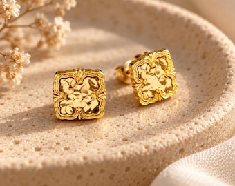 Puede incluir: Un par de pendientes de botón cuadrados de oro con un patrón floral texturizado dentro de un borde decorativo. Los pendientes tienen un acabado pulido y reflectante, diseñados con forma de trébol de cuatro hojas y un intrincado trabajo en metal.