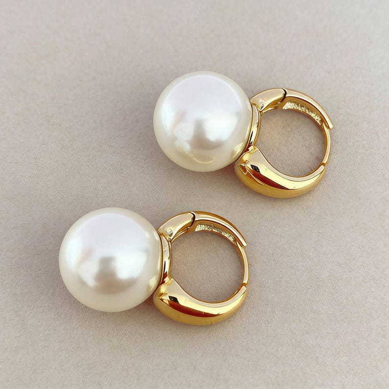 Puede incluir: Un par de pendientes de aro con grandes perlas blancas redondas unidas a aros de metal dorado pulido. Los pendientes cuentan con un mecanismo de cierre de bisagra y muestran una superficie lisa y reflectante tanto en las perlas como en el metal.