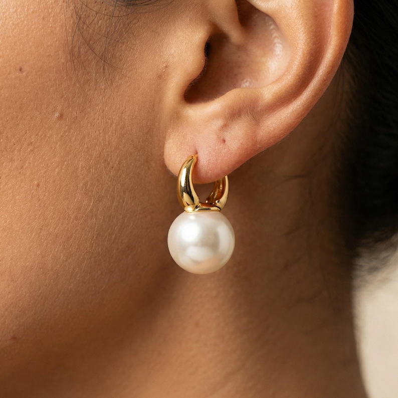 Puede incluir: Un pendiente de aro dorado con acabado liso y pulido, que presenta una gran perla blanca redonda como colgante. El pendiente est&aacute; dise&ntilde;ado con una pieza superior de metal dorado curvado que se conecta a la perla esf&eacute;rica central.