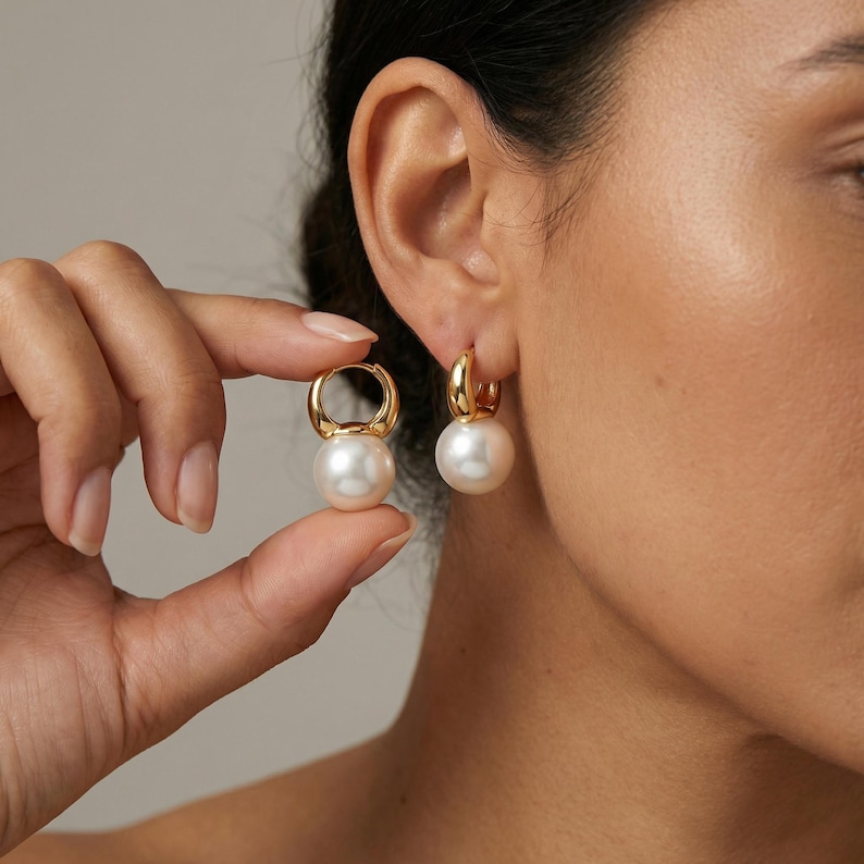 Puede incluir: Un par de pendientes de aro dorados con un acabado liso y pulido. Cada aro est&aacute; adornado con una perla blanca redonda que cuelga de la parte inferior de la banda met&aacute;lica circular, creando un dise&ntilde;o de joyer&iacute;a cl&aacute;sico y equilibrado.