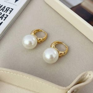 Puede incluir: Un par de pendientes de aro en tono dorado, cada uno con una perla esf&eacute;rica blanca, grande y lisa, suspendida en la parte inferior. Los aros tienen un acabado met&aacute;lico pulido y un mecanismo de cierre con bisagra para el l&oacute;bulo.