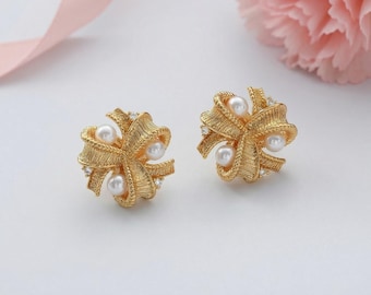 Puede incluir: Un par de pendientes dorados con forma de lazo, con perlas incrustadas y pequeños cristales transparentes. Los pendientes están sobre una superficie blanca, con una cinta rosa y una flor rosa en el fondo.