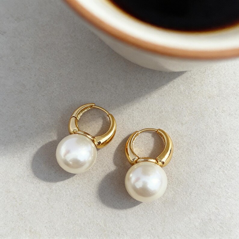 Puede incluir: Un par de pendientes de aro dorados, cada uno con una perla blanca redonda, lisa y &uacute;nica suspendida en la parte inferior. Los aros tienen un acabado met&aacute;lico pulido y las perlas son esf&eacute;ricas y brillantes, creando un dise&ntilde;o de joyer&iacute;a cl&aacute;sico.