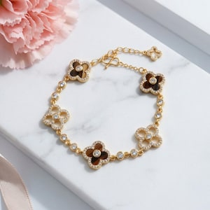 Puede incluir: Pulsera dorada con abalorios en forma de trébol con centros marrones y detalles brillantes. La pulsera tiene una cadena delicada con un cierre de palanca y se muestra sobre una superficie de mármol blanco.