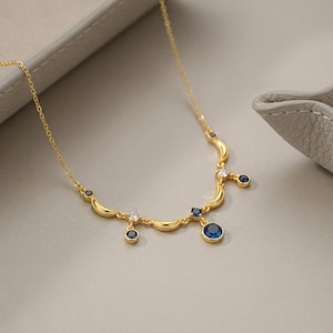 May include: A gold-tone necklace featuring a delicate chain with crescent moon-shaped links, accented by small round blue and clear white stones. The design includes several dangling stone charms suspended from the crescent moon elements.