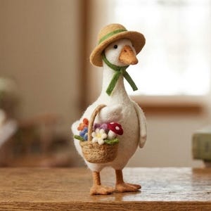May include: A needle-felted white goose figurine wearing a tan straw-style hat with a green ribbon tie. The goose holds a small woven basket filled with colorful felt flowers and a single red and white spotted mushroom in its wing.