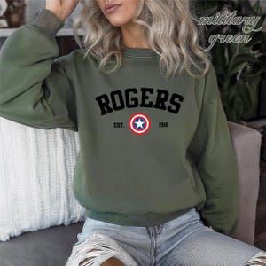 May include: Military green sweatshirt with the word "ROGERS" in black block letters. Below the word "ROGERS" is a circular emblem with a white star in the center, surrounded by a red and blue circle. The text "EST. 1918" is below the emblem.