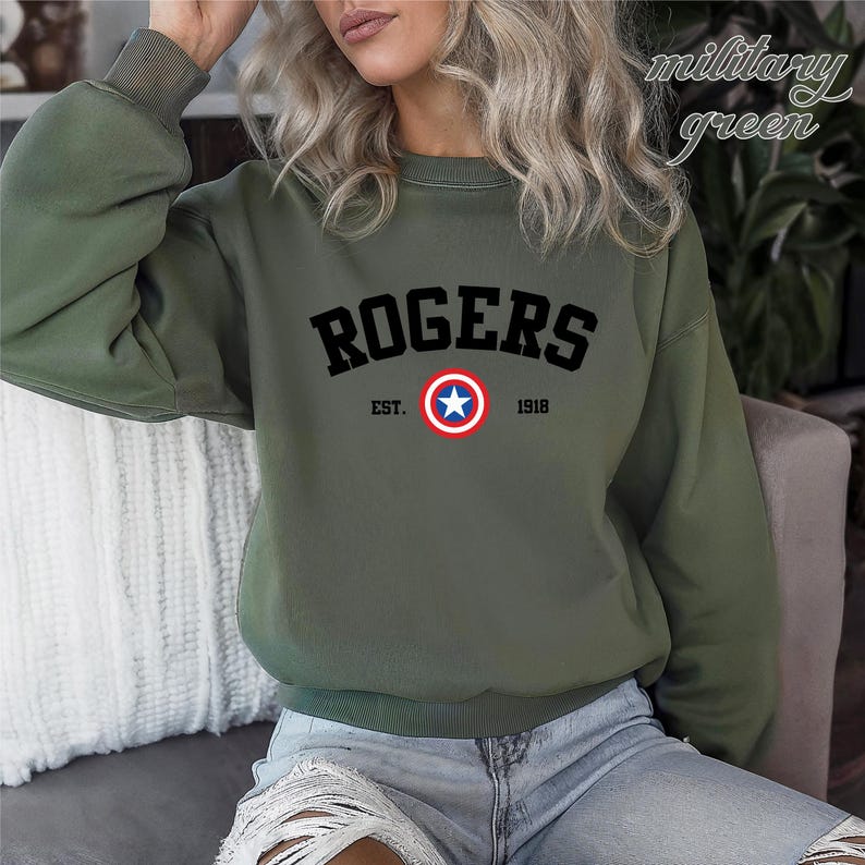 May include: Military green sweatshirt with the word "ROGERS" in black block letters. Below the word "ROGERS" is a circular emblem with a white star in the center, surrounded by a red and blue circle. The text "EST. 1918" is below the emblem.