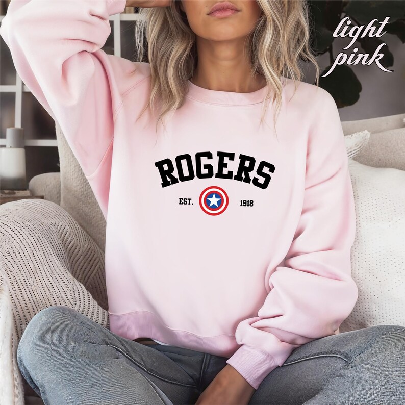 May include: Light pink sweatshirt with the word "ROGERS" in black block letters. Below the word "ROGERS" is a red, white, and blue star emblem, with the text "EST. 1918". The sweatshirt is a crew neck style.