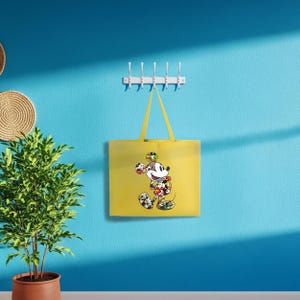 May include: A yellow tote bag with a Mickey Mouse design, hanging on a white coat rack. The bag is against a blue wall. A potted plant and woven wall decor are also visible in the image.