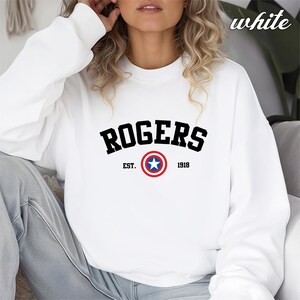 May include: White crewneck sweatshirt with the word "ROGERS" in black block letters. Below the word is a circular emblem with a blue star in the center, surrounded by red and white rings. The text "EST. 1918" is below the emblem.