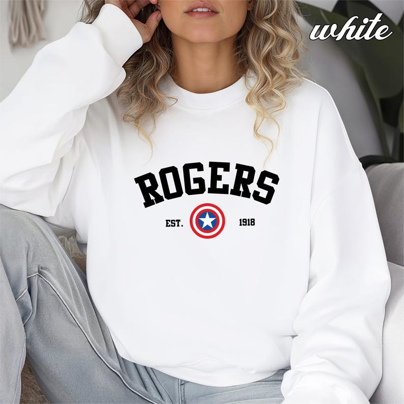 May include: White crewneck sweatshirt with the word "ROGERS" in black block letters. Below the word is a circular emblem with a blue star in the center, surrounded by red and white rings. The text "EST. 1918" is below the emblem.
