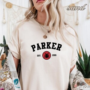 May include: A white t-shirt with black text that reads "Parker Est. 2001" with a red spider graphic in the center.