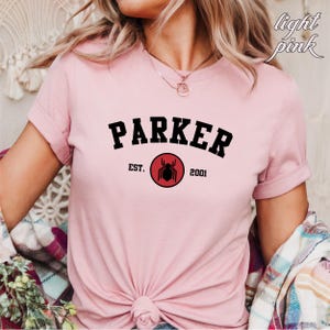 May include: Pink t-shirt with black text that reads "Parker Est. 2001" and a red circle with a black spider graphic.