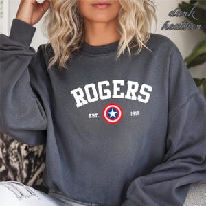 May include: Dark heather gray sweatshirt with the word "ROGERS" in white block letters. Below the word is a circular emblem with a blue star in the center, surrounded by red and white rings. The text "EST. 1918" is below the emblem.