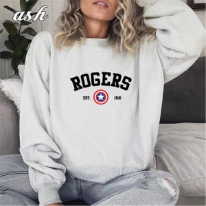 May include: A light gray sweatshirt with the word "ROGERS" in black block letters. Below the word is a circular emblem with a blue star in the center, surrounded by a red and white ring. The text "EST. 1918" is below the emblem.