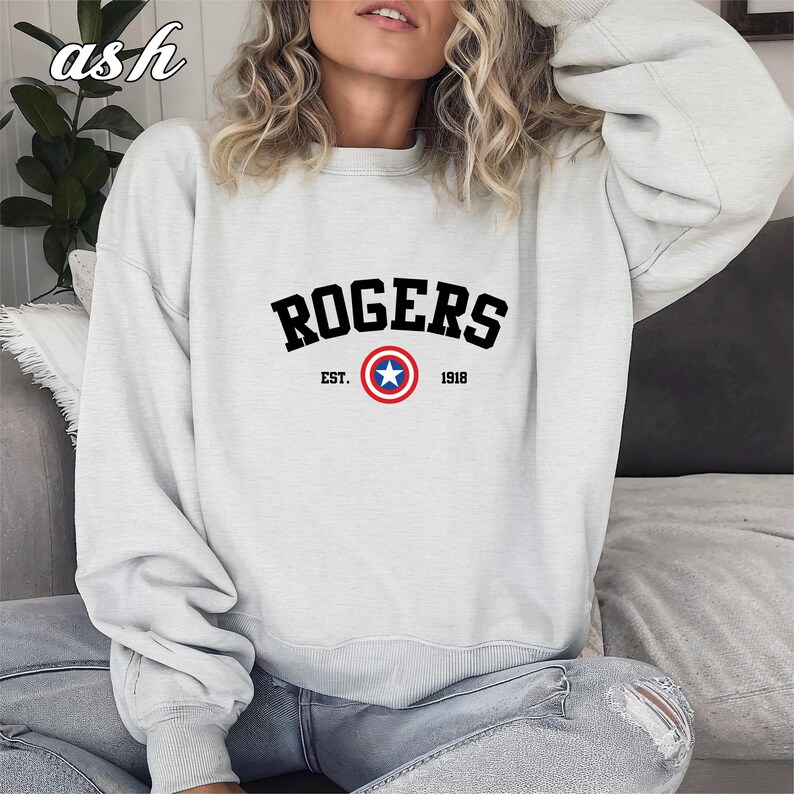 May include: A light gray sweatshirt with the word "ROGERS" in black block letters. Below the word is a circular emblem with a blue star in the center, surrounded by a red and white ring. The text "EST. 1918" is below the emblem.