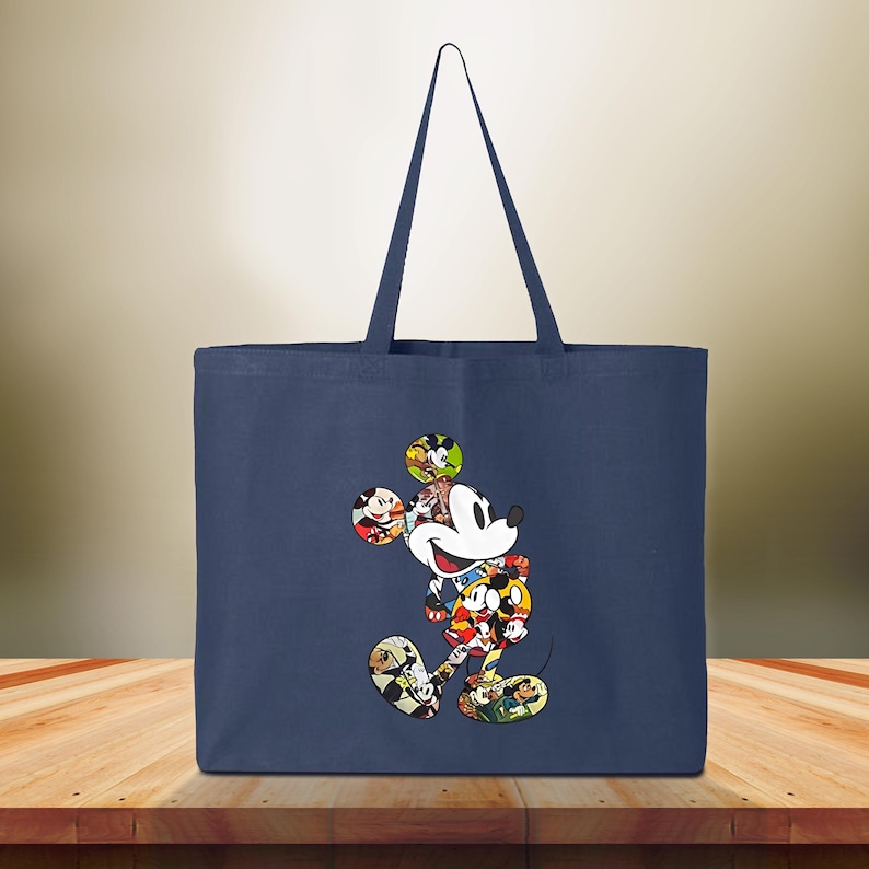 May include: Navy blue tote bag with a colourful Mickey Mouse design. The cartoon character is made up of various images of Mickey Mouse. The bag has two handles and is made of a sturdy fabric. The background is a blurred wooden surface.