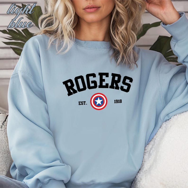 May include: Light blue sweatshirt with the word "ROGERS" in black arched letters. Below the word "ROGERS" is the text "EST. 1918" and a circular emblem with a white star and red and blue rings.