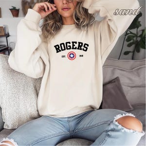 May include: A sand-colored sweatshirt with the word "ROGERS" in bold black letters across the chest. Below the name is "EST. 1918" and a circular emblem with a blue star. The sweatshirt is long-sleeved and has a crew neck.