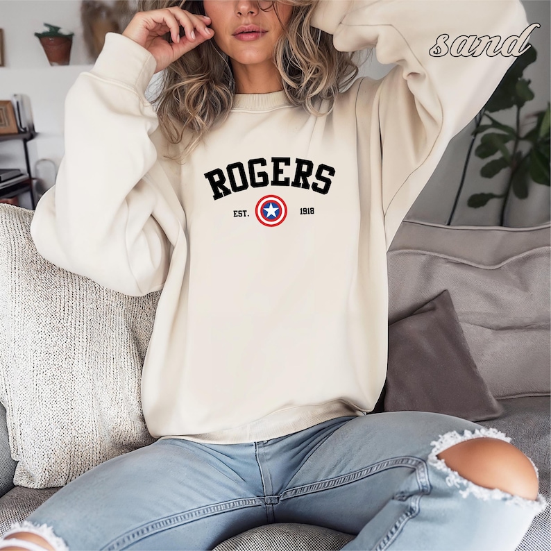 May include: A sand-colored sweatshirt with the word "ROGERS" in bold black letters across the chest. Below the name is "EST. 1918" and a circular emblem with a blue star. The sweatshirt is long-sleeved and has a crew neck.