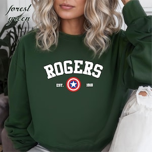 May include: Forest green sweatshirt with the word "ROGERS" in white block letters. Below the word "ROGERS" is a circular emblem with a blue star in the center, surrounded by a red and white ring. The text "EST. 1918" is below the emblem.