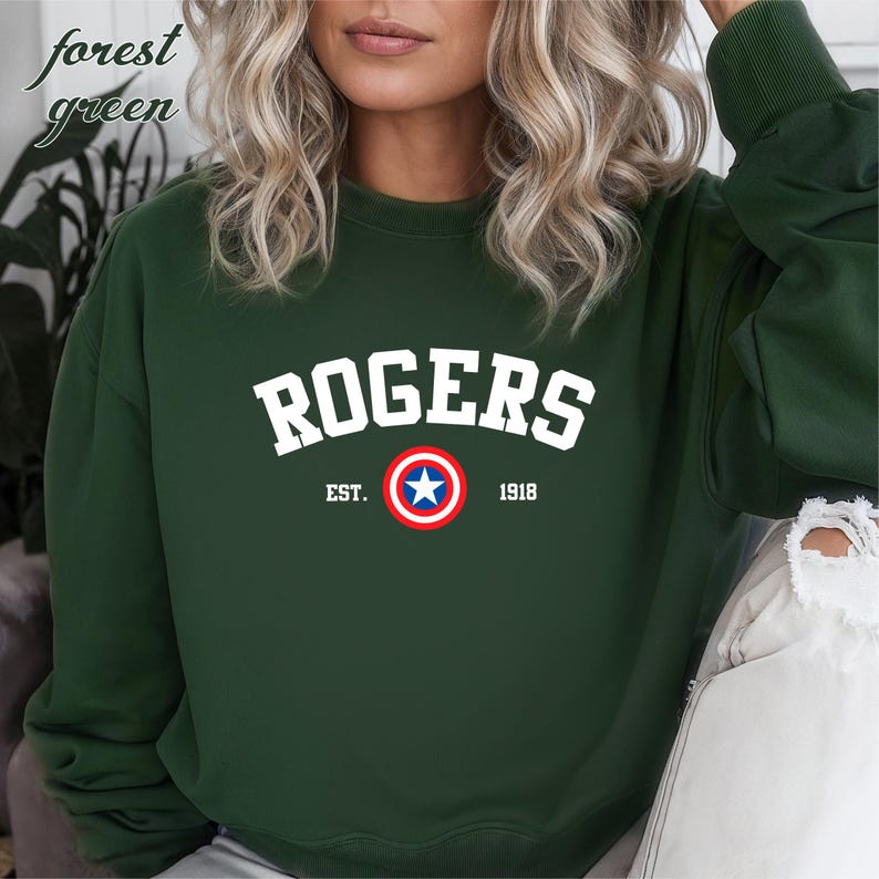 May include: Forest green sweatshirt with the word "ROGERS" in white block letters. Below the word "ROGERS" is a circular emblem with a blue star in the center, surrounded by a red and white ring. The text "EST. 1918" is below the emblem.