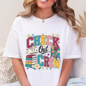 May include: A white short-sleeve t-shirt featuring a colorful graphic print with the text "CHECK Out CREW". The design includes a smiling library card, a stack of books, a rolling book cart, and daisy flowers in shades of red, yellow, blue, and pink.
