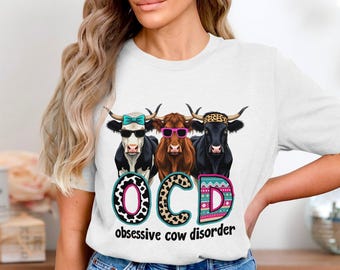 May include: White t-shirt featuring three cartoon cows wearing sunglasses and a headband. The text "OCD obsessive cow disorder" is printed below the cows in a colorful design. The cows are black and white, brown, and black.