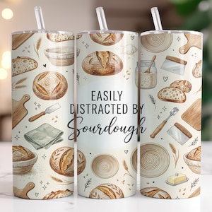May include: A stainless steel tumbler with a white background and a repeating pattern of bread, baking tools, and wheat. The text "EASILY DISTRACTED BY Sourdough" is centered on the tumbler.