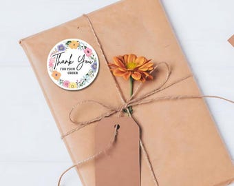 Può includere: Un pacco di carta marrone legato con spago e un piccolo fiore. Un adesivo rotondo con un bordo floreale dice "Grazie per il tuo ordine".