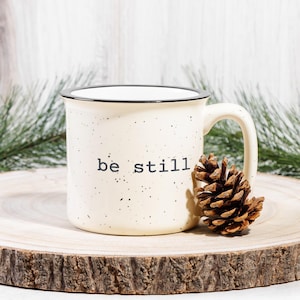 May include: Cream-colored enamel mug with black speckles and a black rim. The words "be still" are printed in black. A pine cone rests beside the mug on a wooden slice. Green pine branches are in the background.